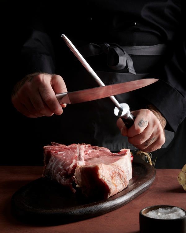 Chef sharpening kitchen knife with honing steel over raw steak on cutting board