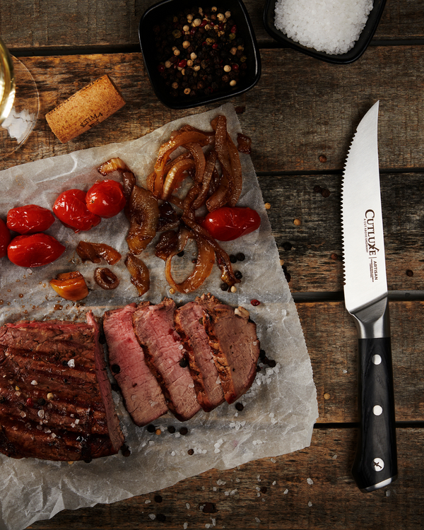 Serrated kitchen knife beside sliced steak, roasted tomatoes, caramelized onions, salt, and peppercorns on rustic wood