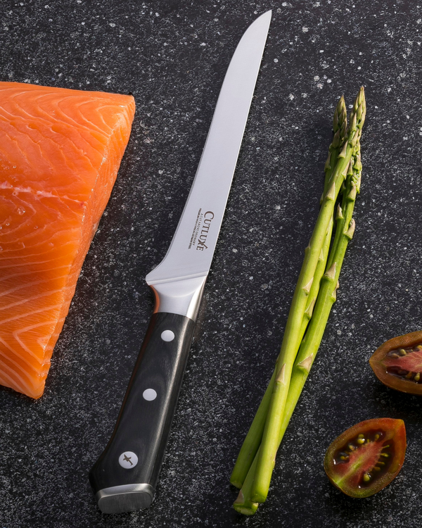 Sharp kitchen knife with salmon, asparagus, and tomato on dark surface
